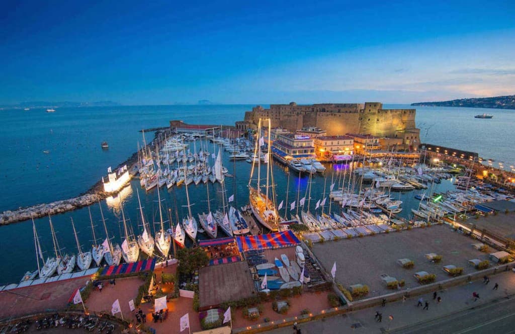 Borgo Marinari e Castel dell’Ovo a Napoli al crepuscolo: porticciolo con barche a vela, luci calde sui locali sul mare e cielo blu sul Golfo.